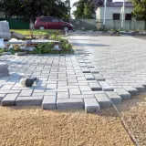 Trwa utwardzenie terenu pod parking. Układanie kostki brukowej, w tle samochód i materiały budowlane.