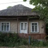 Stary dom z lat 80. z falistym dachem i drewnianą werandą, otoczony bujną zielenią.