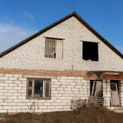 Niedokończony dom z białej cegły, z pustymi oknami i zniszczonym wejściem, wygląda jak po częściowym zawaleniu się budynku.