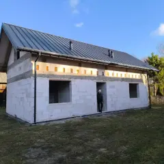 Budowa domu bez pozwolenia w toku. Białe bloczki, szary dach, puste okna. Ktoś stoi w drzwiach.