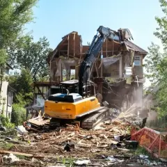 Żółty koparka prowadzi rozbiórkę starego domu, zasypując teren gruzem.