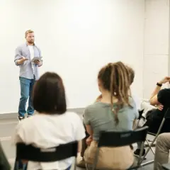 Mężczyzna przemawia do grupy osób trzecich na zebraniu wspólnoty. Słuchacze siedzą na krzesłach, jeden z nich opiera głowę na dłoni.