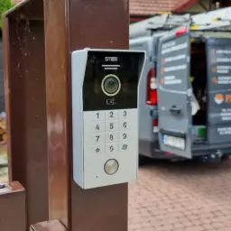 Nowoczesny domofon z kamerą i klawiaturą numeryczną zamontowany na słupku bramy. W tle widać zaparkowany samochód dostawczy.