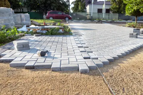 Trwa utwardzenie terenu pod parking. Układanie kostki brukowej, w tle samochód i materiały budowlane.