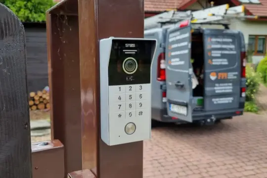 Nowoczesny domofon z kamerą i klawiaturą numeryczną zamontowany na słupku bramy. W tle widać zaparkowany samochód dostawczy.