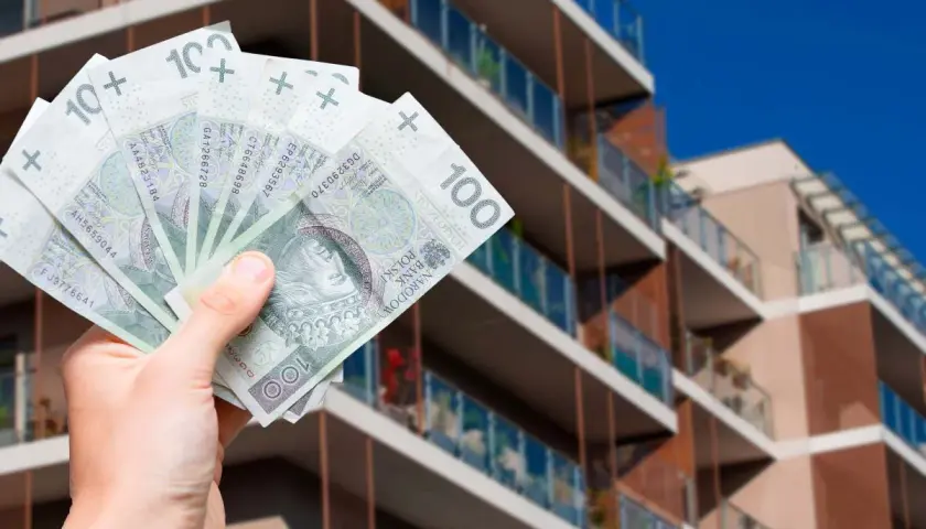 Dłoń trzyma banknoty 100 zł na tle nowoczesnego apartamentowca. Pytanie: jak zapłacić podatek od wynajmu mieszkania?