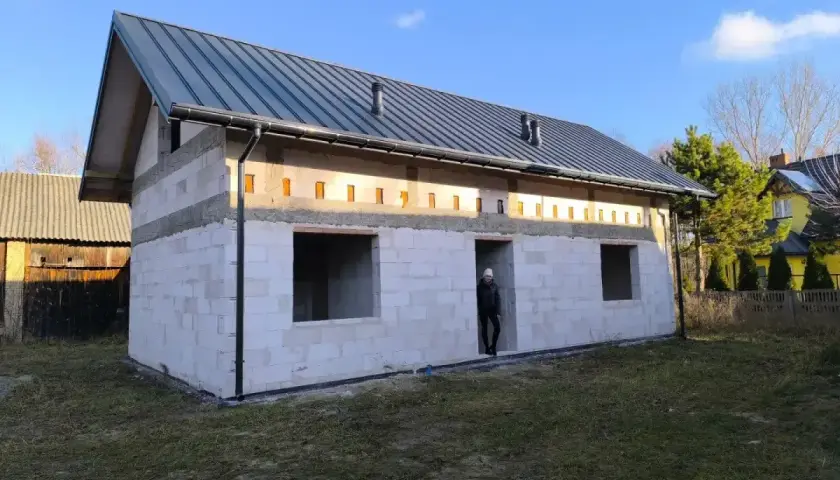 Budowa domu bez pozwolenia w toku. Białe bloczki, szary dach, puste okna. Ktoś stoi w drzwiach.