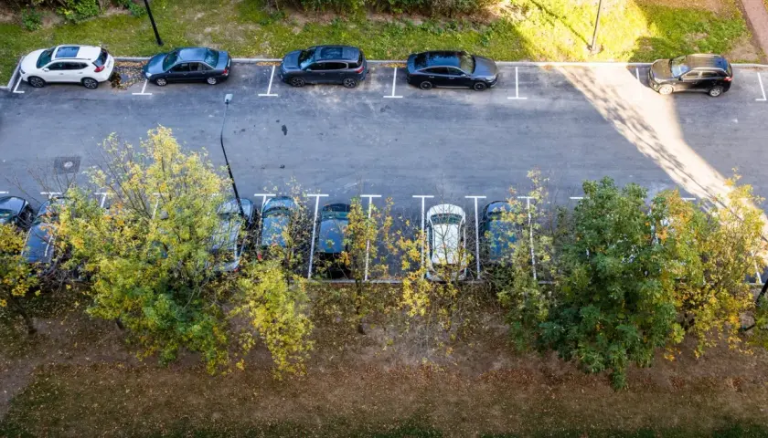 Widok z góry na parkowanie samochodów dostawczych na osiedlu. Kilka aut stoi na wyznaczonych miejscach, otoczonych zielenią i drzewami.