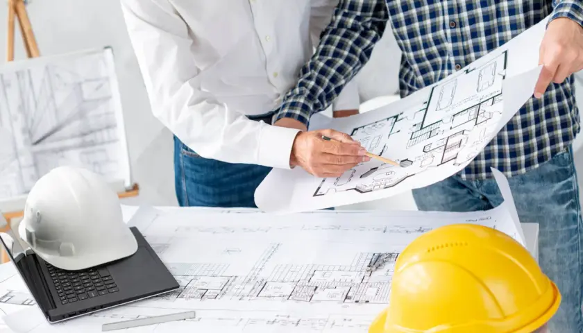 Architekci analizują plany, omawiając podział wspólnoty mieszkaniowej. Na stole leżą kaski i laptop.