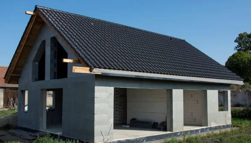 Dom w stanie surowym, z betonowych pustaków, z czarnym dachem i otwartymi otworami okiennymi i drzwiowymi.