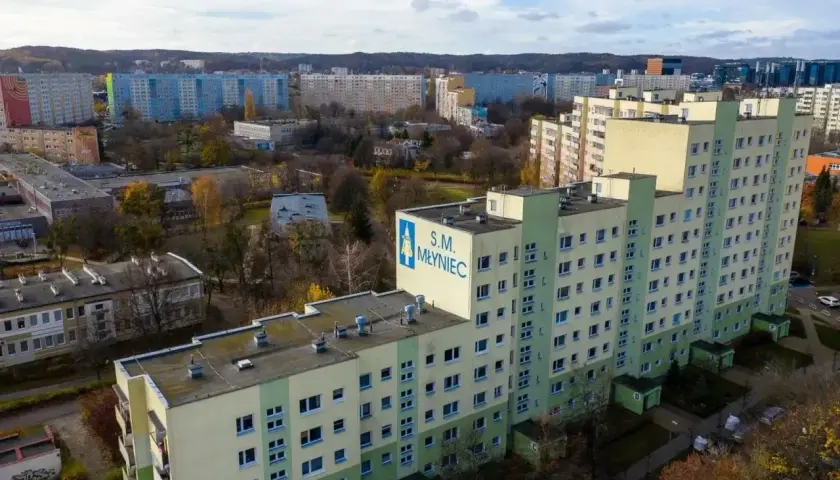 Duży blok mieszkalny S.M. Młyniec z charakterystycznym logo, otoczony innymi budynkami i drzewami jesienią.