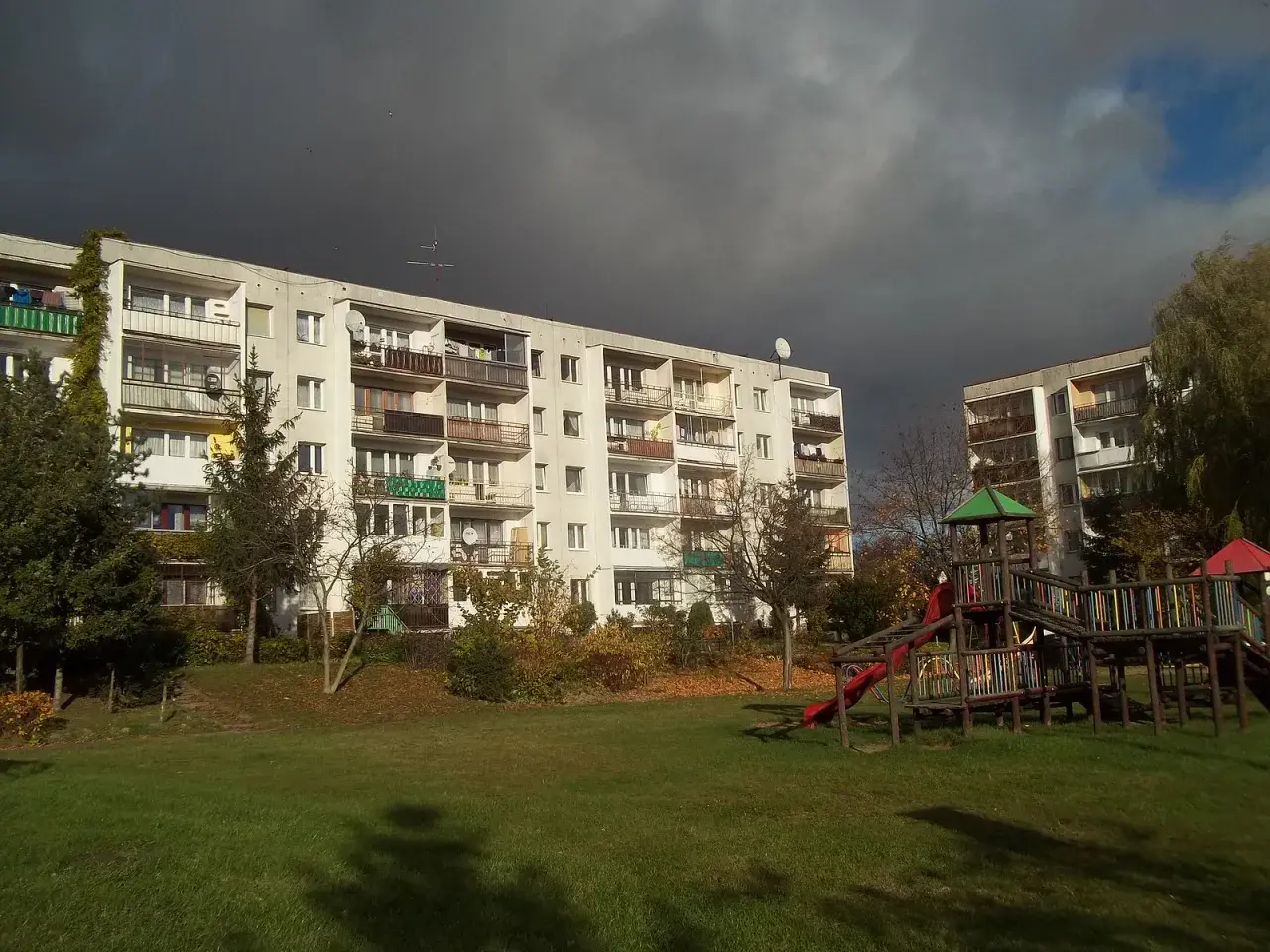 Osiedle z wielkiej płyty, plac zabaw i jesienne drzewa pod burzowym niebem.