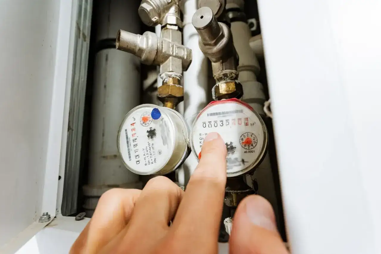 Dłoń wskazuje na licznik ciepła podczas montażu. Widać dwa liczniki i metalowe rury.