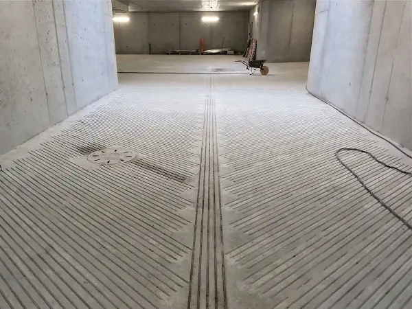 Betonowa pochylnia do garażu podziemnego z antypoślizgowymi nacięciami. Widać studzienkę kanalizacyjną i wózek widłowy w oddali.