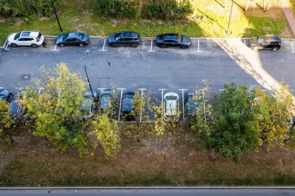 Widok z góry na parkowanie samochodów dostawczych na osiedlu. Kilka aut stoi na wyznaczonych miejscach, otoczonych zielenią i drzewami.